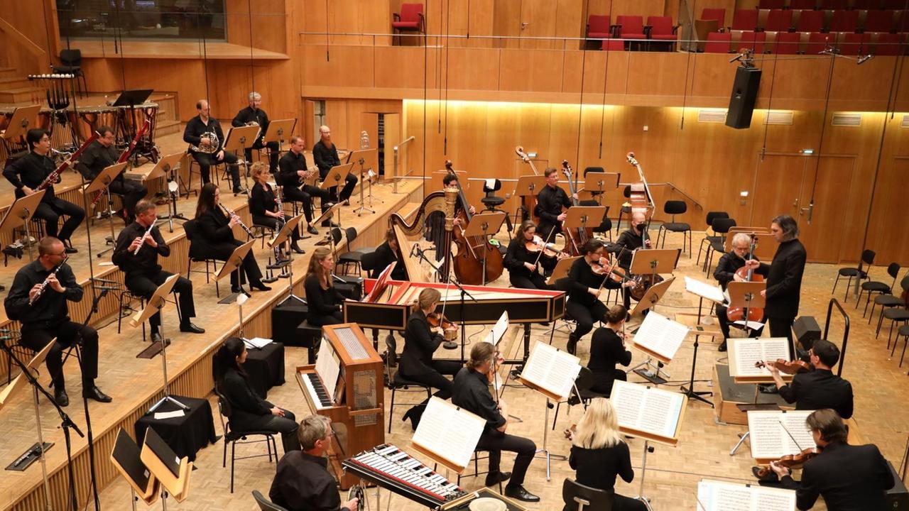 Orchestermusiker mit Cembalo sitzten in größere Abständen voneinander und spielen nach Weisungen des Dirigenten.