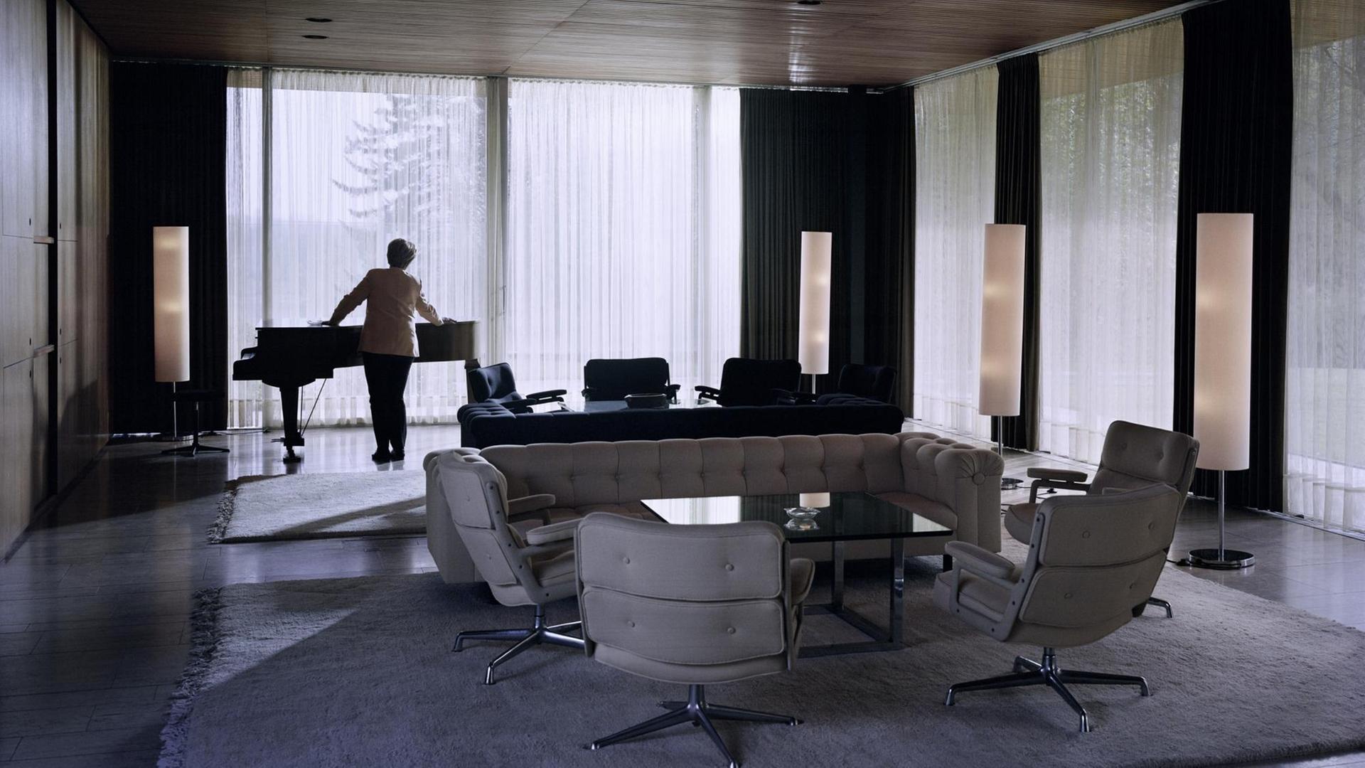 Eine Doppelgängerin von Angela Merkel steht an einem Flügel im Kanzlerbungalow.