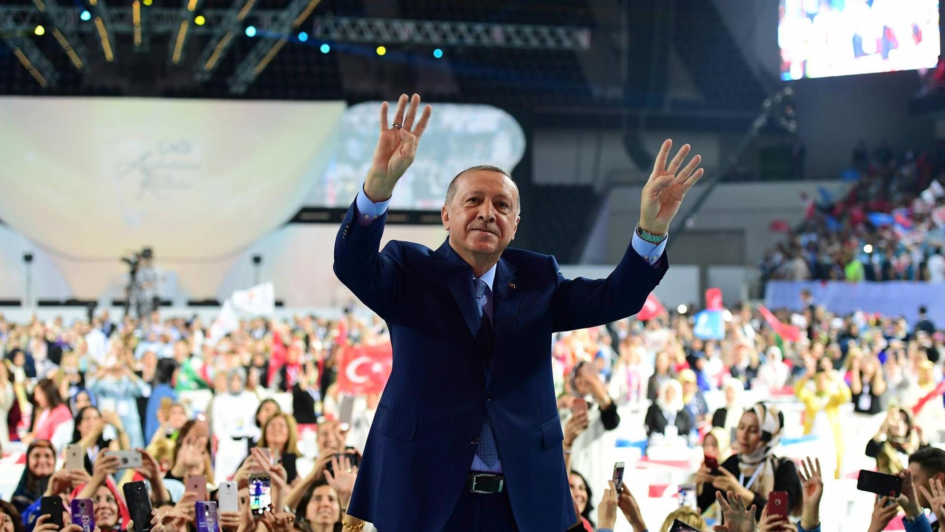 Der türkische Präsident Erdogan steht bei einer Veranstaltung seiner Partei AKP mit dem Rücken zum Publikum und hebt die Hände zum Rabia-Gruß nach oben. Dabei zeigen jeweils vier Finger nach oben.