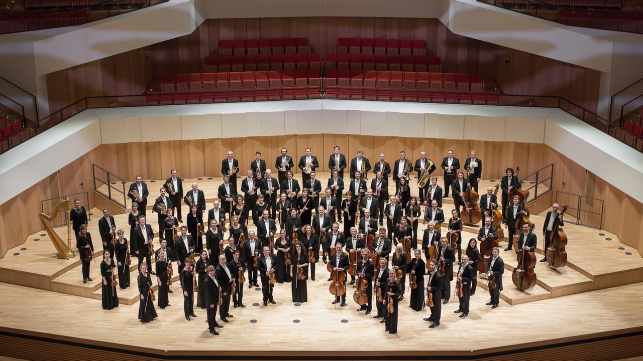 Die Mitglieder eines Orchesters stehen in Konzertkleidung auf der Bühne eines Konzertsaals.