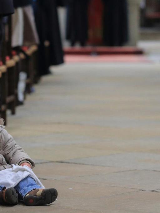 Ein kleines Mädchen mit Schnuller und Zöpfen sitzt neben der Bank in einer vollbesetzten Kirche.