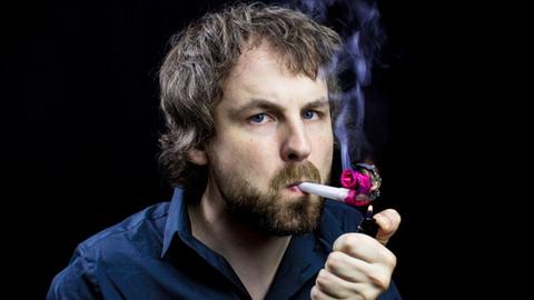Portrait von Tino Bomelino, der anstatt einer Zigarette eine Papiertröte im Mundwinkel hat und sich diese mit einem Feuerzeug ansteckt.