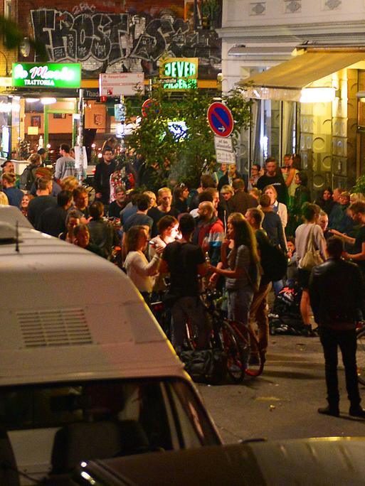 Junge Erwachsene stehen abends in einem Ausgehviertel auf der Straße und trinken.