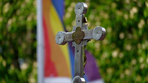 Ein Kreuz steht bei einem Open-Air-Segnungsgottesdienst für Liebende vor einer Kirche. Im Hintergrund ist eine Regenbogenfahne zu sehen.