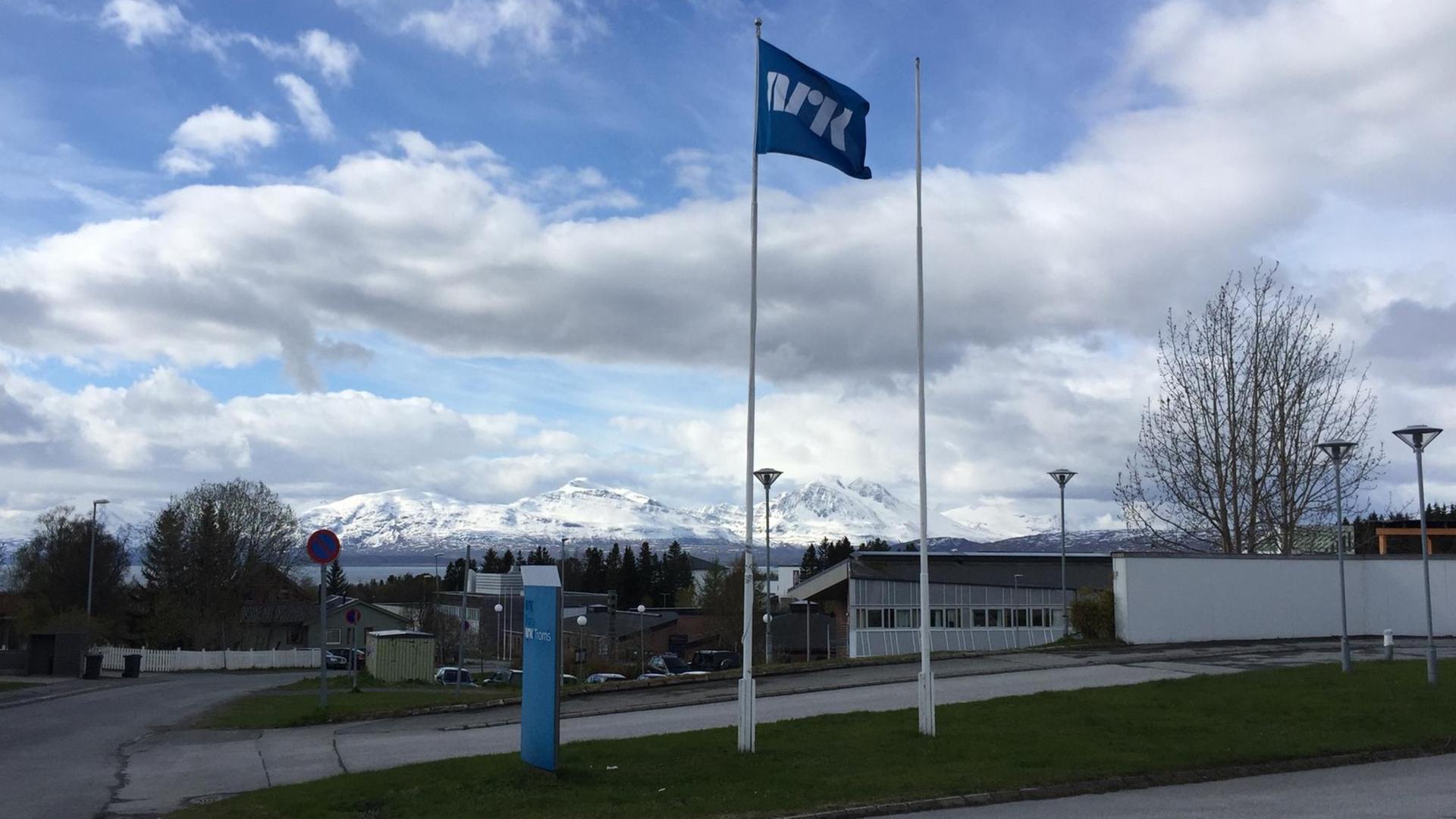 Zu sehen ist eine Fahne vor dem Redaktionsgebäude des norwegischen Rundfunksenders NRK in Tromsø. Im Hintergrund sind schneebedeckte Berge zu erkennen.