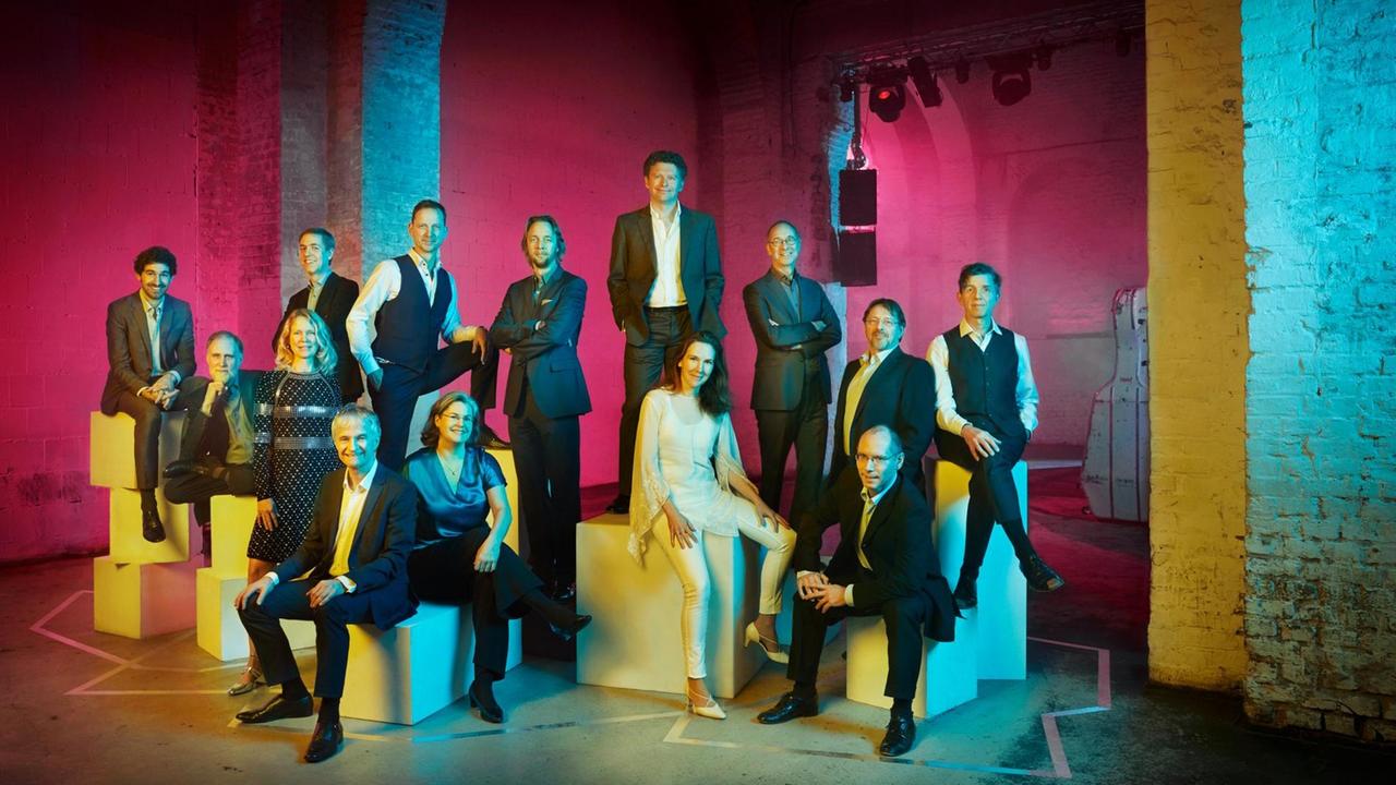 Die Musiker des Ensembles stehen und sitzen in einer bunt beleuchteten Ecke eines Betonbaus mit unterschiedlichen Sitzquadern.