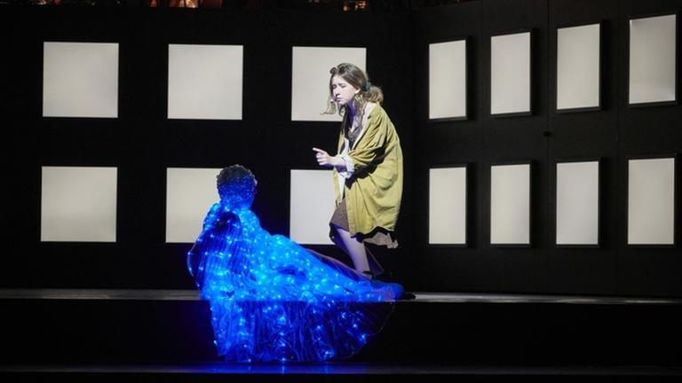 Ein junger Mann mit einem Kostüm aus blauen LED-Lichtern sitzt auf der Bühne, neben ihm eine junge Frau