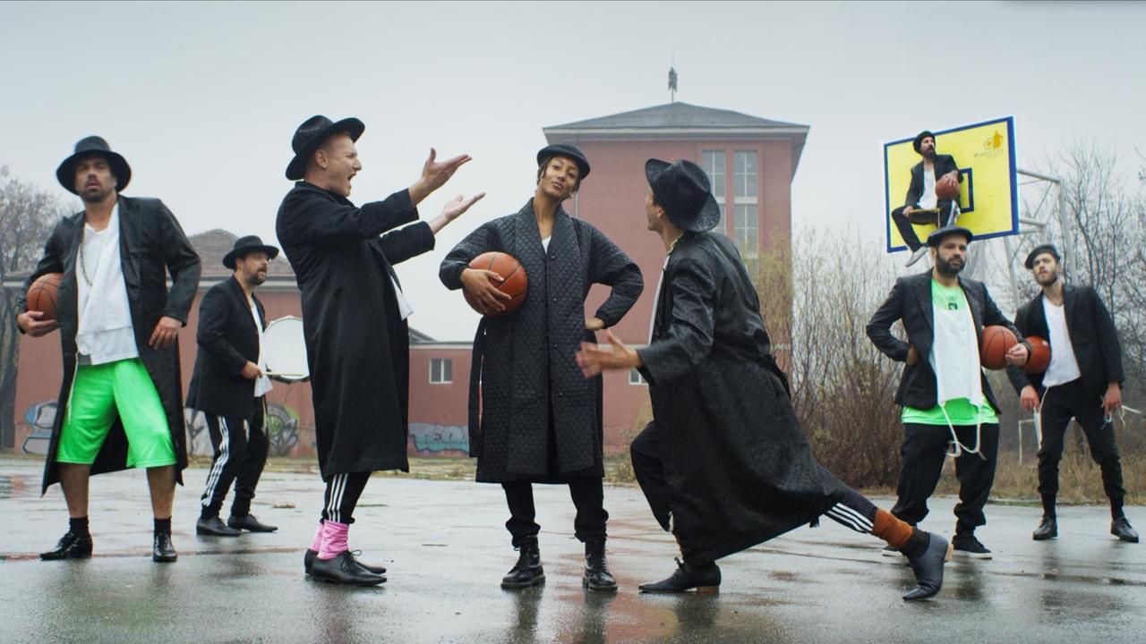 Die Bandmitglieder inszenieren sich auf einem Basketballplatz. Alle tragen einen Hut, einige einen Basketball unter dem Arm.
