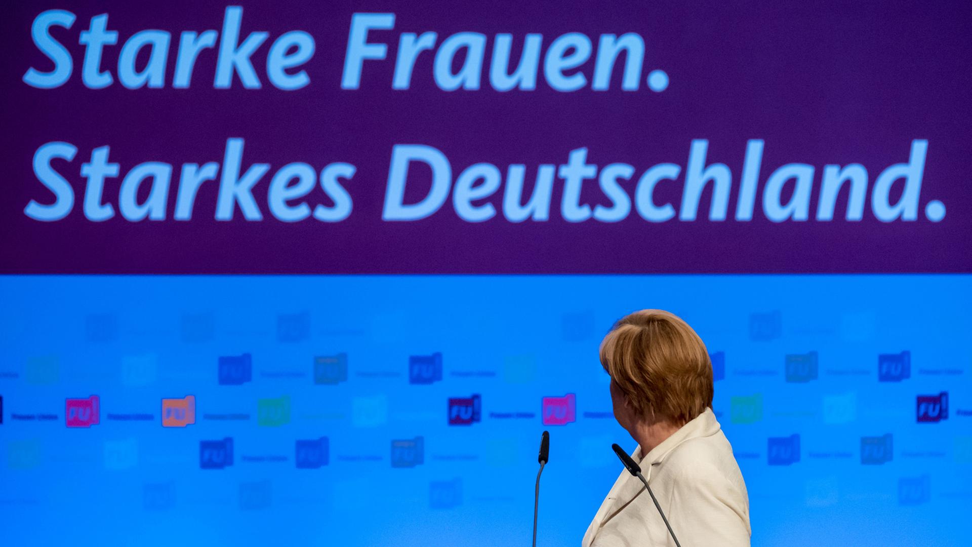 Bundeskanzlerin Merkel am Rednerpult, sie hat sich umgedreht und schaut auf das Banner "Starke Frauen. Starkes Deutschland" hinter sich.