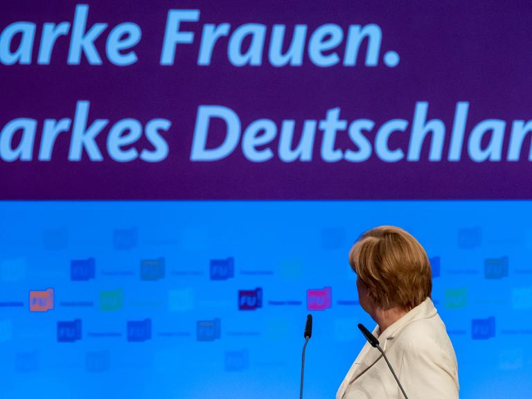 Bundeskanzlerin Merkel am Rednerpult, sie hat sich umgedreht und schaut auf das Banner "Starke Frauen. Starkes Deutschland" hinter sich.