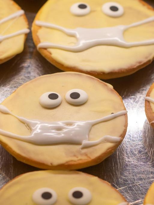 Kleine runde Kuchen in einer Auslage: Sie sind mit Zuckerguss verziert. Ihnen wurden zwei Augen und ein Mundschutz "aufgemalt".