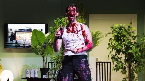 Ein Schauspieler im Unterhemd bespritzt sich mit einer blutroten Flüssigkeit aus einer Flasche.