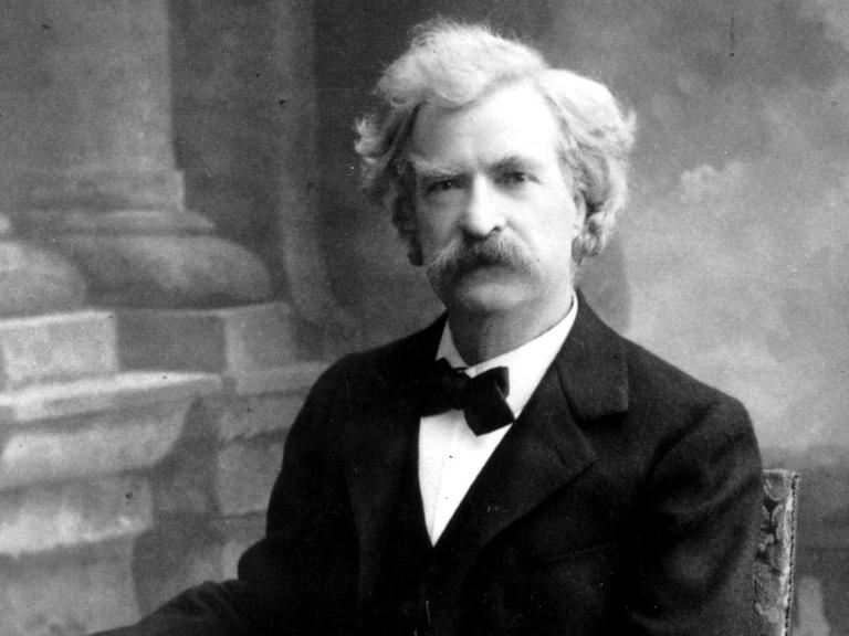 Mark Twain sitzt in einem dunklen Anzug auf einem Stuhl.