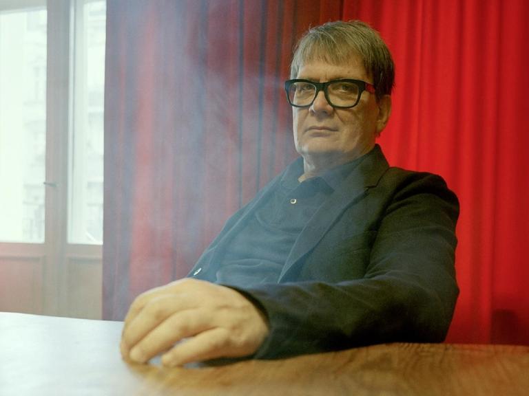 Sven Regener trägt eine schwarze Brille und einen schwarzen Anzug und sitzt an einem Holztisch vor einem Fenster mit roten Vorhängen. Es steigt Rauch von unter dem Tisch auf.