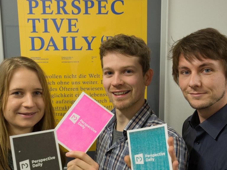 Die Medienmacher Maren Urner, Bernhard Eickenberg (r) und Han Langeslag halten am 04.02.2016 in Münster Werbepostkarten mit dem Namen ihres geplanten Online-Magazins «Perspective Daily» vor einem gleichnamigen Werbeplakat in den Händen.