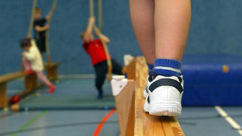 Ein Kind balanciert auf einer Bank, im Hintergrund hangeln andere Kinder während des Kinderturnens an Seilen.