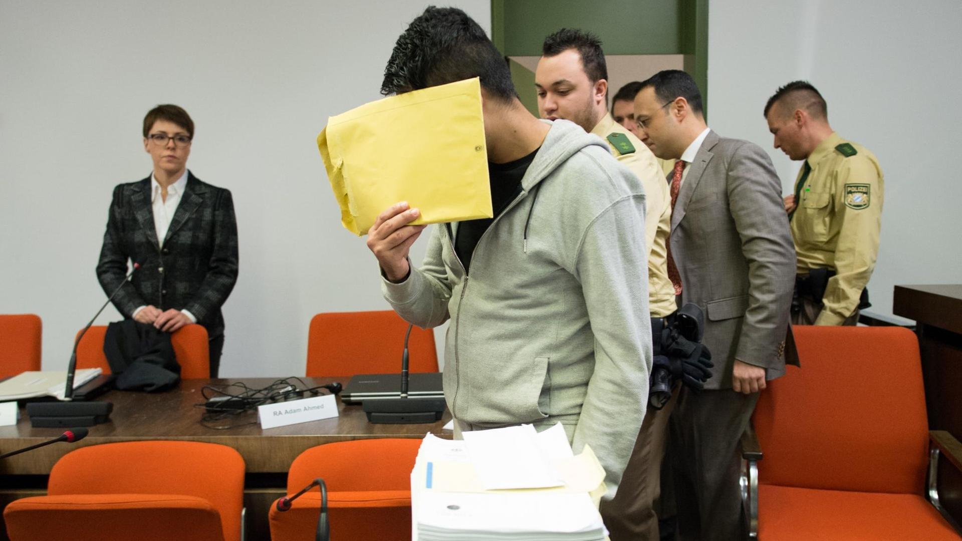 Ein junger Mann versteckt als Angeklagter am 20.01.2015 in einem Gerichtssaal sein Gesicht hinter einem Umschlag. Um ihn herum stehen Polizisten und weitere Personen.