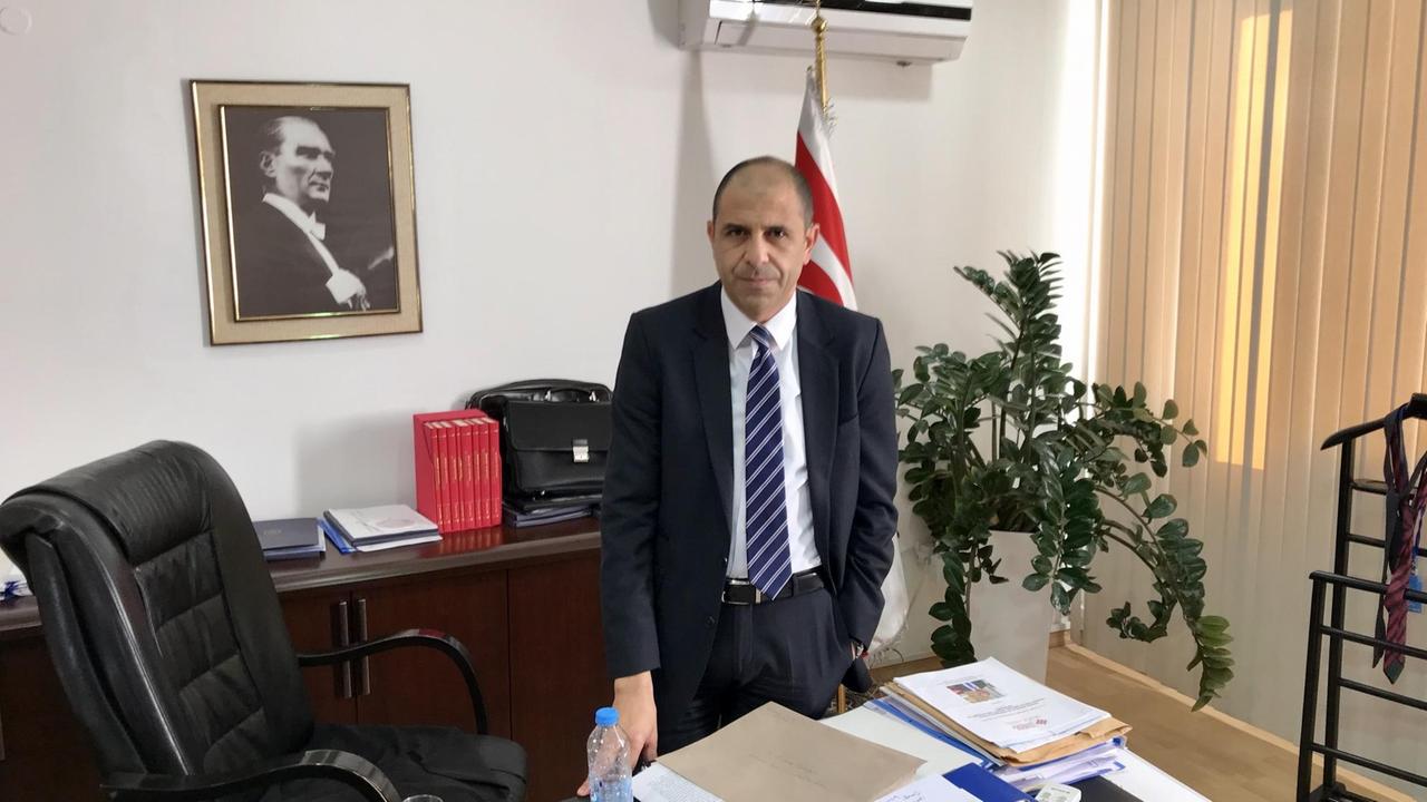 Ein Mann mit Halbglatze gekleidet in Anzug und Krawatte steht in seinem Büro vor dem Schreibtisch, hinter ihm hängt eine gerahmte Fotografie von Atatürk.
