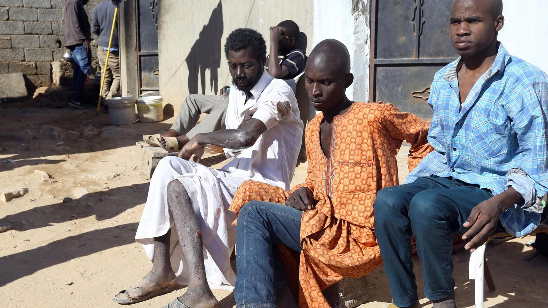 Männer aus afrikanischen Staaten sitzen an einer Häuserwand auf einer unbefestigten Straße.
