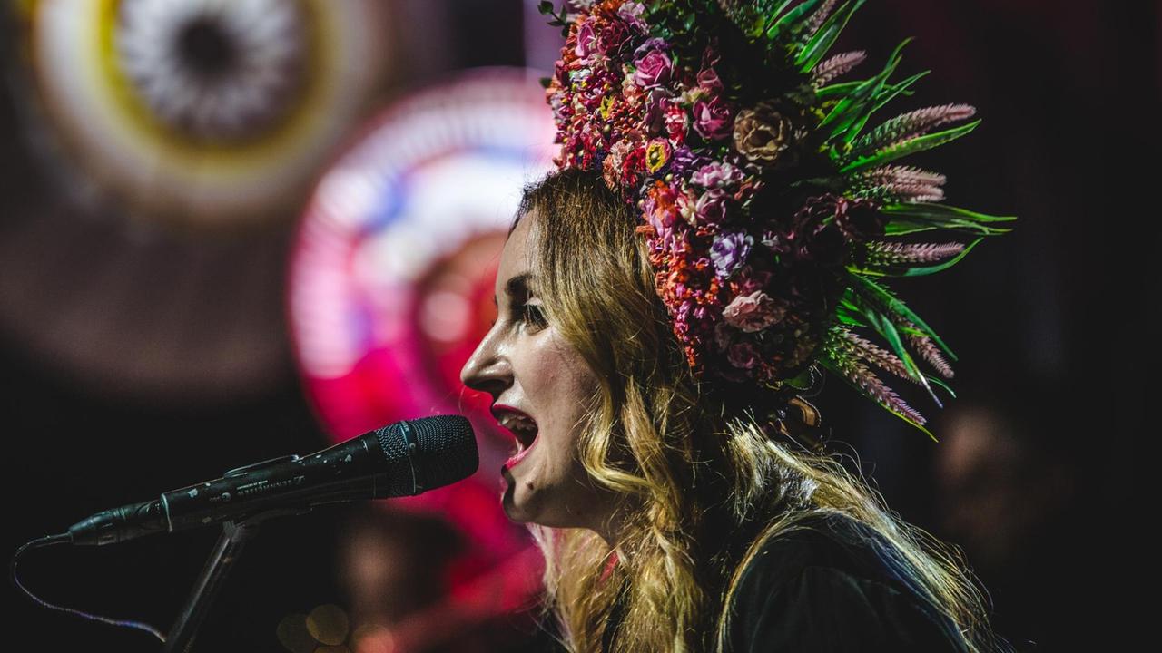 Eine Frau mit einem bunten Hut aus Blumen sitzt vor einem Mikrofon und singt.