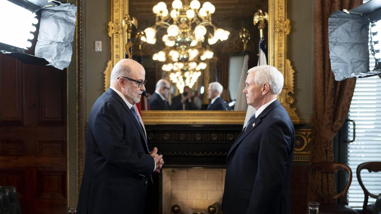 Zwei Männer (Ex-US-Vizepräsident Mike Pence und Journalist Mark Levin) stehen sich in einem herrschaftlichen Raum gegenüber. Im Hintergrund hängt ein Spiegel, in dem sich ein Kronleuchter spiegelt.