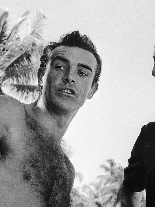 Schwarzweißfoto von Sean Connery mit nacktem Oberkörper und dem Schriftsteller Ian Fleming unter freiem Himmel.