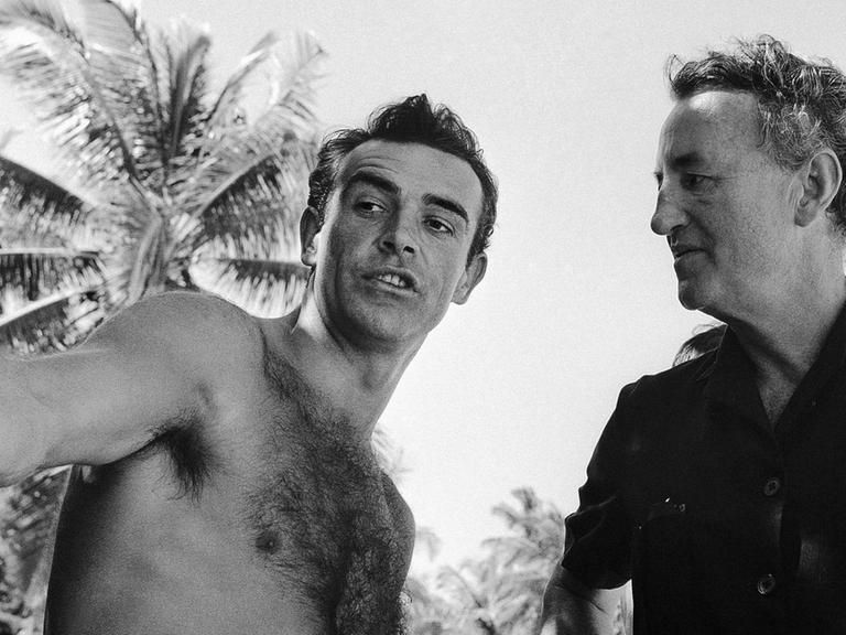 Schwarzweißfoto von Sean Connery mit nacktem Oberkörper und dem Schriftsteller Ian Fleming unter freiem Himmel.