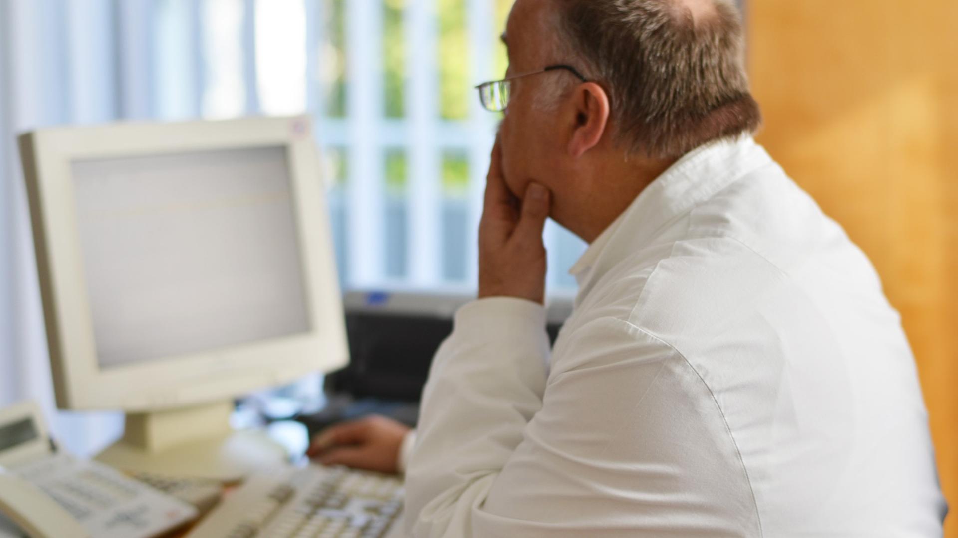 Ein Hausarzt sitzt in seinem Beratungszimmer vor einem Computer
