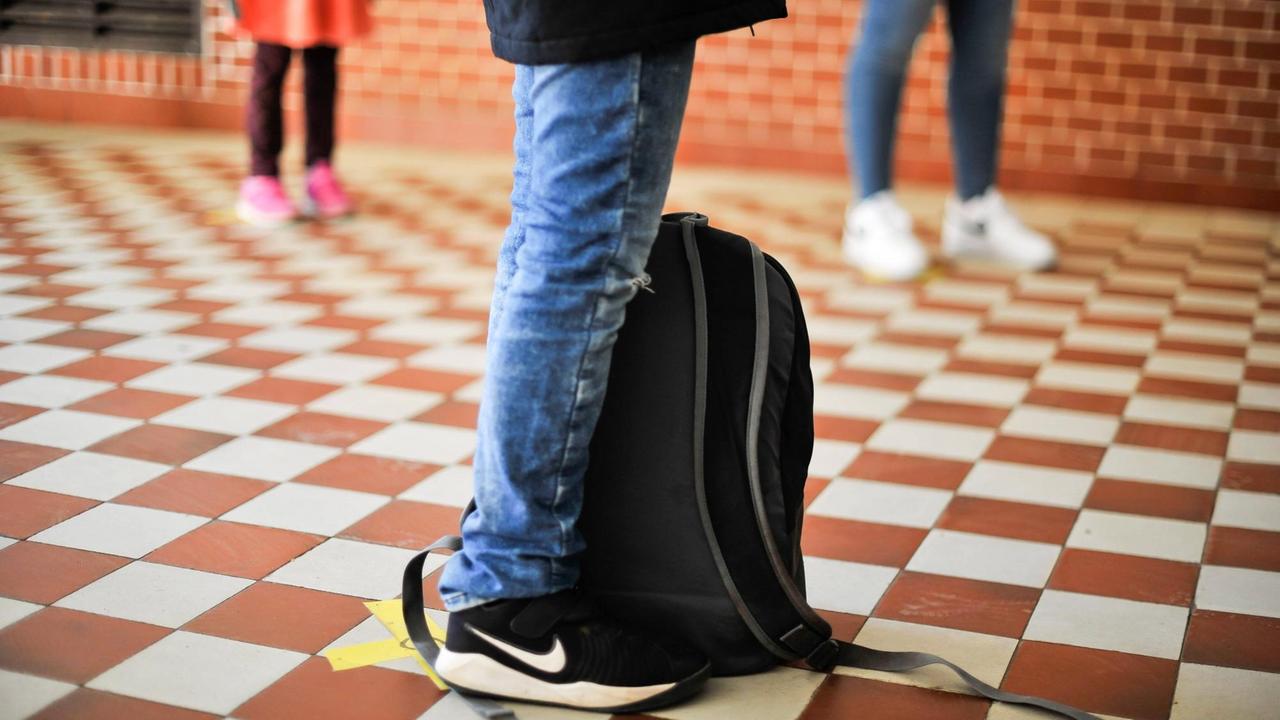 Schülerbeine in Jeans und Turnschuhen stehen mit Abstand voneinander im Flur einer Schule.