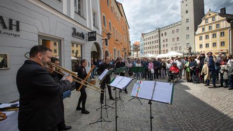 Vier Musiker und eine Musikerin in schwarzer Konzertkleidung spielen mit Blasinstrumenten in einer Fußgängerzone. Vor ihnen stehen Notenständer, dahinter zahlreiche Menschen, die zuhören.