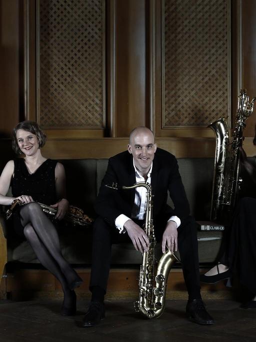 Vier Musiker sitzen mit ihren unterschiedlich großen Saxophonen in Abendrobe auf einer Polsterbank in einem mit dunklem, schweren Holz vertäfelten Raum und schauen lachend in die Kamera.