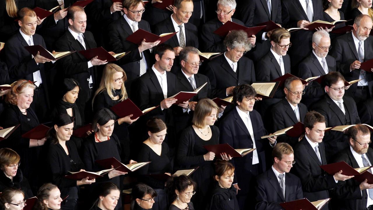 Männer und Frauen in schwarzer Kleidung und mit roten Gesangsbüchern stehen dicht an dicht in mehreren Reihen, die sich diagonal durch das Bild ziehen.