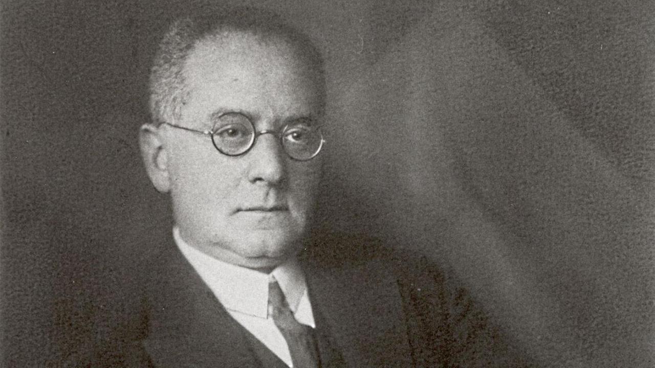 Schwarz-weiß Bild, Carl Flesch sitzend, trägt einen dunklen Anzug mit Weste und Krawatte, eine runde Brille, ein weißes Einstecktuch