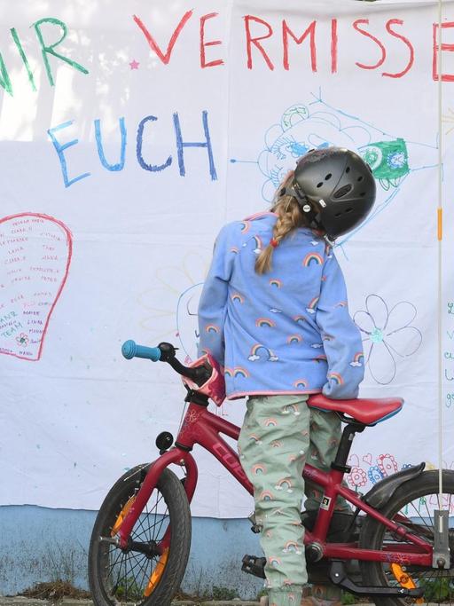 Ein Mädchen auf dem Fahrrad steht vor der Fassade einer Kita mit der Aufschrift "Wir vermissen euch".