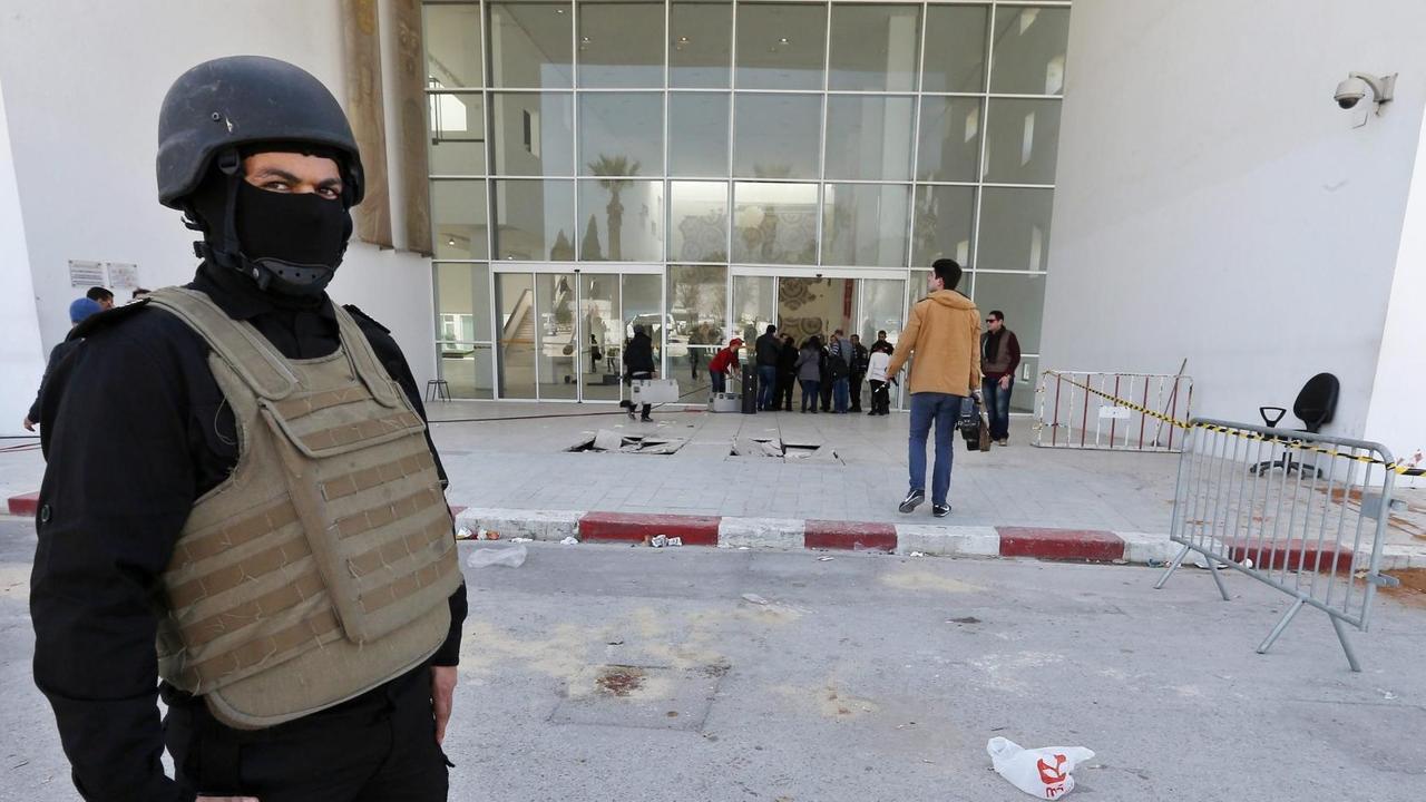 Ein schwarz gekleideter Polizist mit Helm, Sturmhaube und kugelsicherer Weste steht vor dem abgesperrten Eingang des Bardo-Museums in Tunis. Hinter ihm sieht man Ermittler; rechts vom Eingang sind Blutspuren zu sehen. 