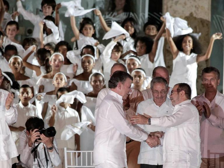 Drei Männer in weißen Händen schütteln sich die Hände, umgeben von vielen anderen Männern und Frauen, die Fahnen schwingen und auch in Weiß gekleidet sind.