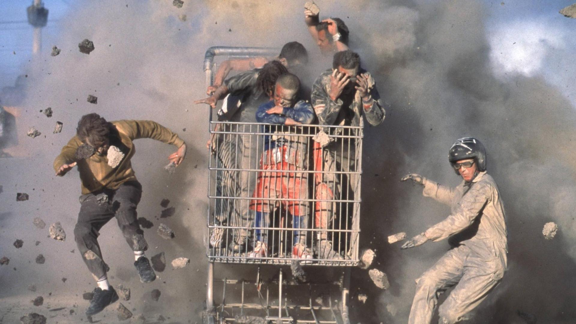 Stuntman fahren in einem vollbesetzten Einkaufswagen, während im Hintergrund etwas explodiert.
