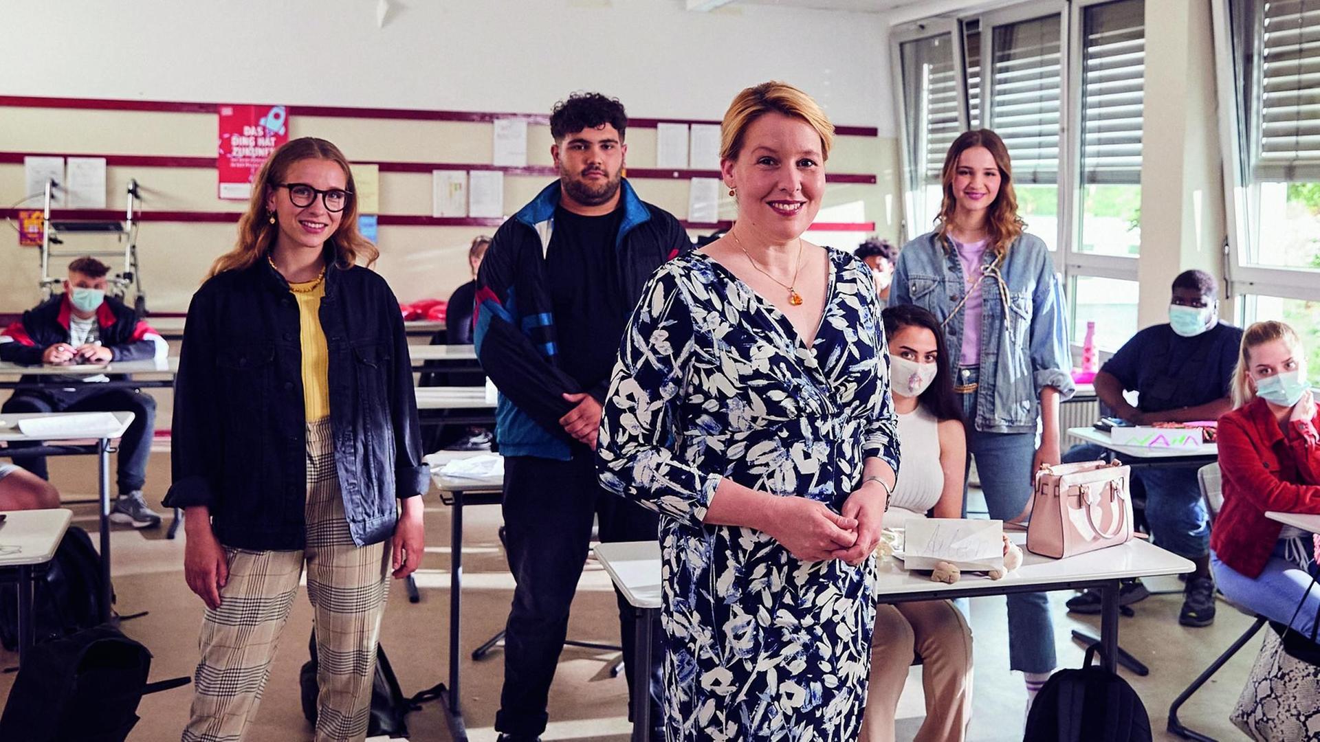 In einem Klassenzimmer stehen die drei Hauptdarsteller der Miniserie "Ehrenpflegas" und Bundesfamilienministerin Franziska Giffey.