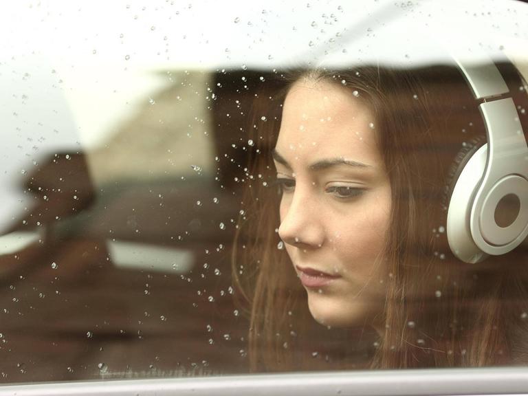 Eine junge Frau mit Kopfhörern blickt gedankenverloren aus einem Autofenster.