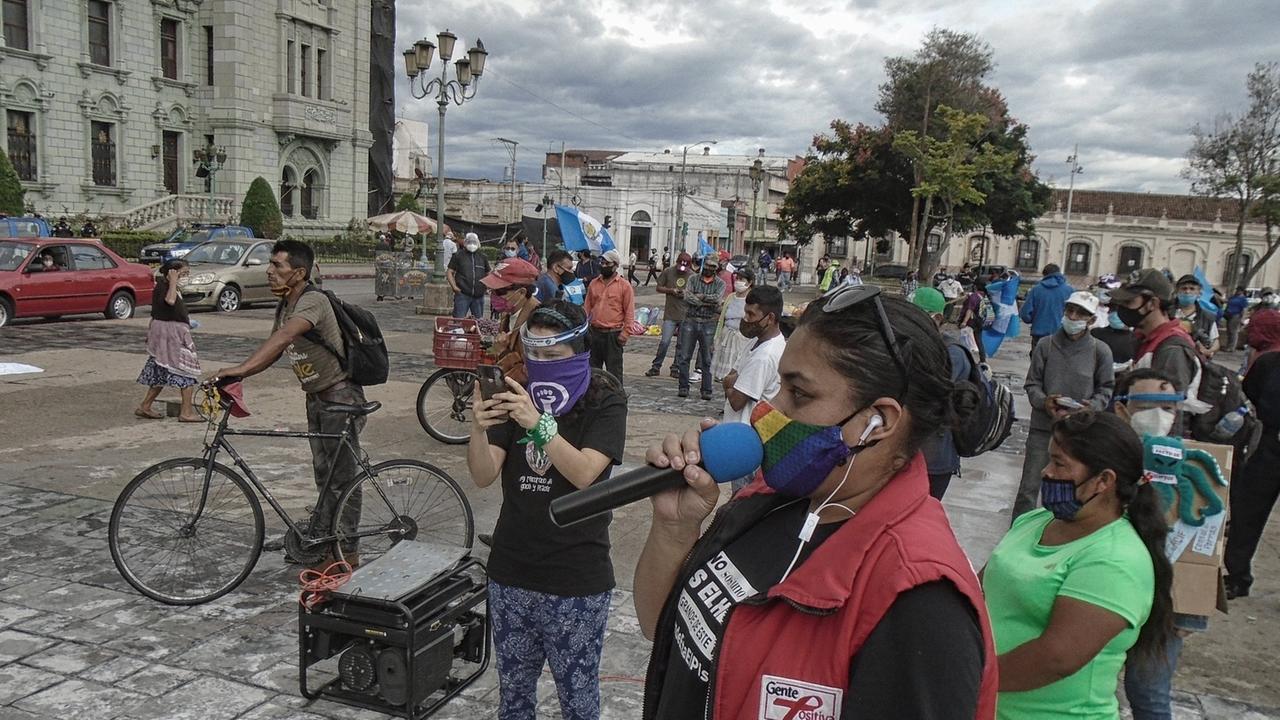 Demonstrierende in Guatemala-Stadt haben sich zum Protest gegen Korruption versammelt, eine Frau in roter Weste spricht in ein Mikrofon, alle tragen Mund-Nase-Masken und halten Abstand voneinander, um sich während der weltweiten Covid-19-Pandemie vor Ansteckung zu schützen.