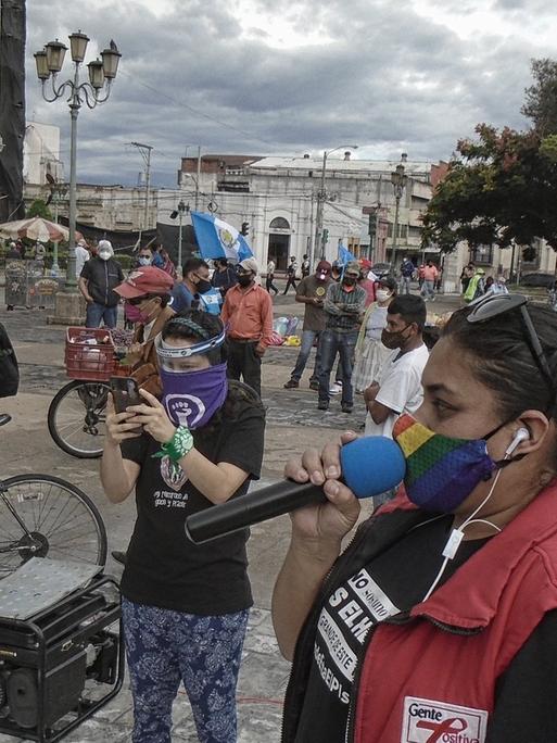Demonstrierende in Guatemala-Stadt haben sich zum Protest gegen Korruption versammelt, eine Frau in roter Weste spricht in ein Mikrofon, alle tragen Mund-Nase-Masken und halten Abstand voneinander, um sich während der weltweiten Covid-19-Pandemie vor Ansteckung zu schützen.