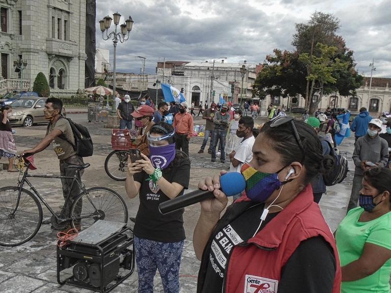 Demonstrierende in Guatemala-Stadt haben sich zum Protest gegen Korruption versammelt, eine Frau in roter Weste spricht in ein Mikrofon, alle tragen Mund-Nase-Masken und halten Abstand voneinander, um sich während der weltweiten Covid-19-Pandemie vor Ansteckung zu schützen.