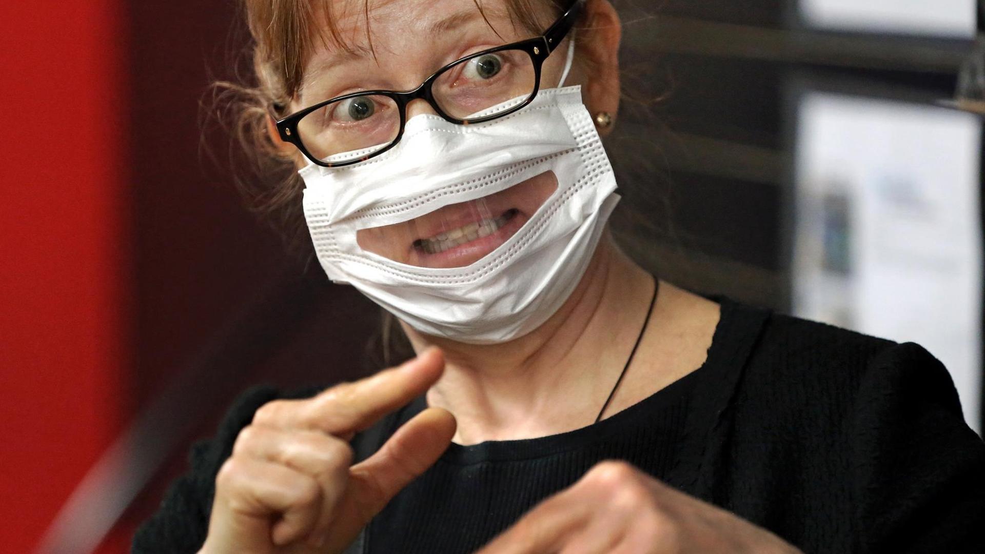 Eine Frau trägt eine Maske mit einem Sichtfenster und macht eine Geste aus der Gebärdensprache