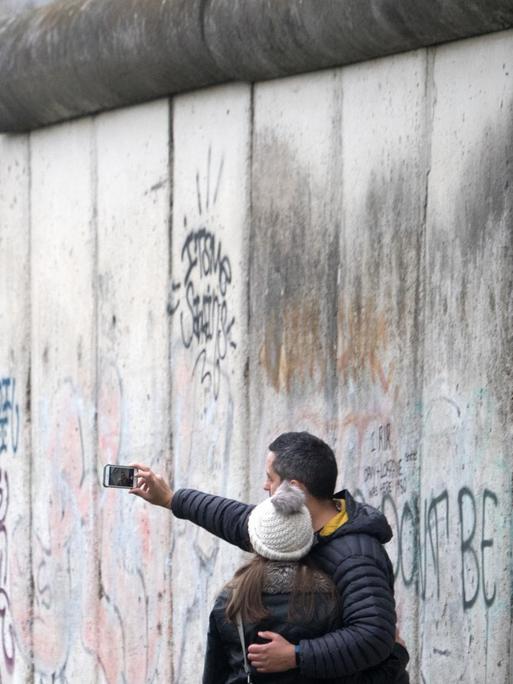 Ein Paar steht vor der Berliner Mauer und macht ein Selfie.