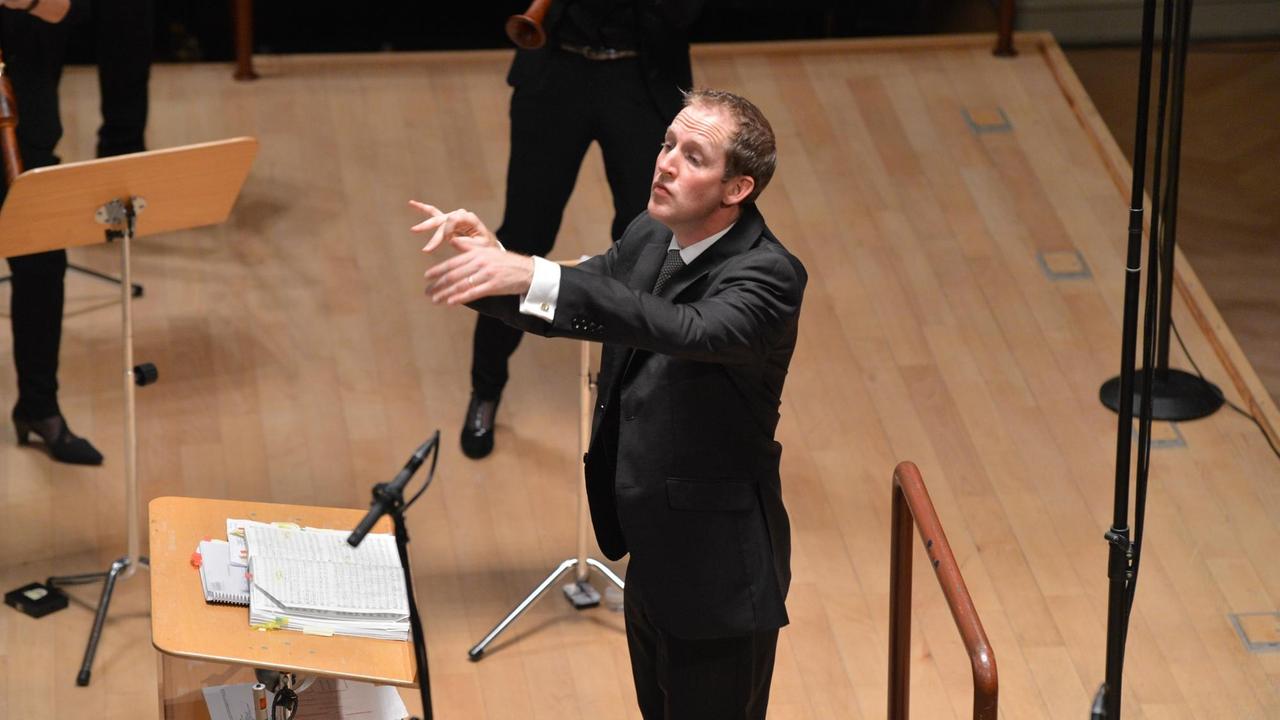 Justin Doyle ist Chefdirigent des RIAS Kammerchors seit Beginn der Saison 2017-18
Er dirigiert mit den Händen, trägt einen schwarzen Anzug, vor ihm steht eine Notenpult und ein Mikrofon