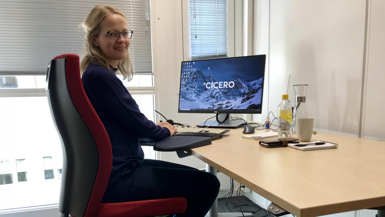 Eine junge blonde Frau mit Brille sitzt an ihrem Schreibtisch, auf dem nur einige Getränke stehen. Ihre Hände liegen auf der Tastatur ihres Computers, auf dessen Bildschirm mit großen Buchstaben "Cicero" steht.