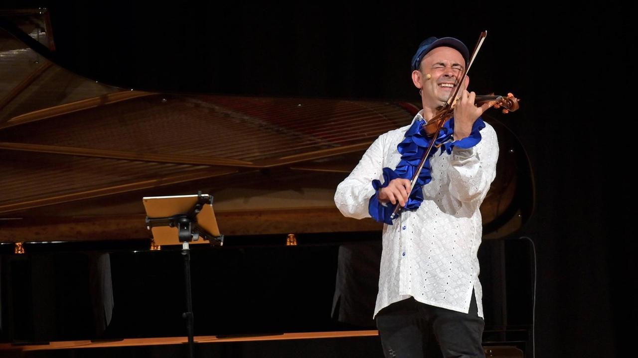 Der russisch-deutschen Geiger, Komponisten, Dirigenten und Schauspieler Aleksey Igudesman in weiß-blauen Hemd und mit Mütze spielt im August 2020 Geige und kneift die Augen dabei zusammen.
