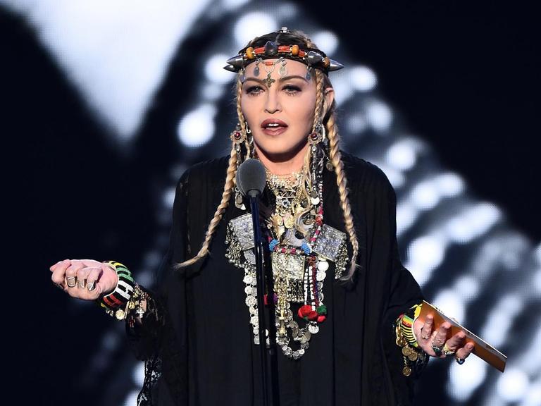 Das Bild zeigt die Sängerin Madonna während einer Rede. Sie trägt ein schwarzes Gewand mit vielen Lagen silbernen Schmuck um ihren Hals und um ihre Handgelenke. Auf dem Kopf trägt sie eine Art Krone mit spitzen Zacken.