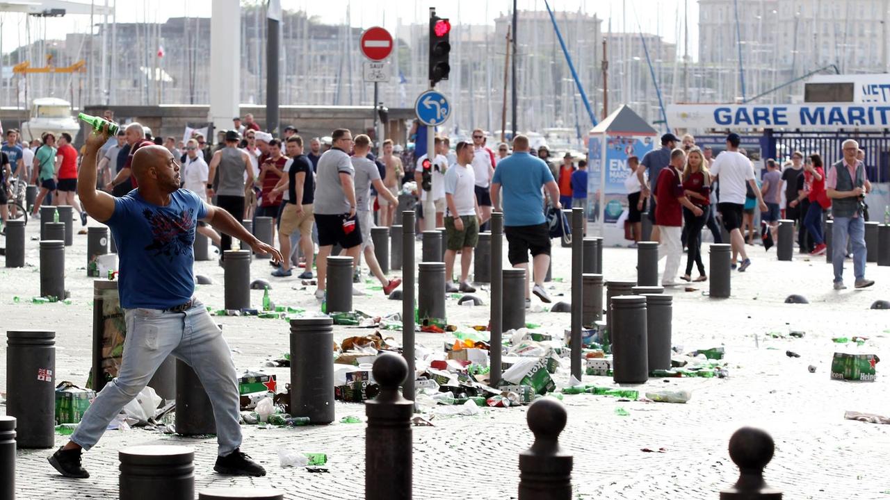Ein Mann wirft eine Bierflasche, auf den Straßen liegt Müll, im Hintergrund Menschen.