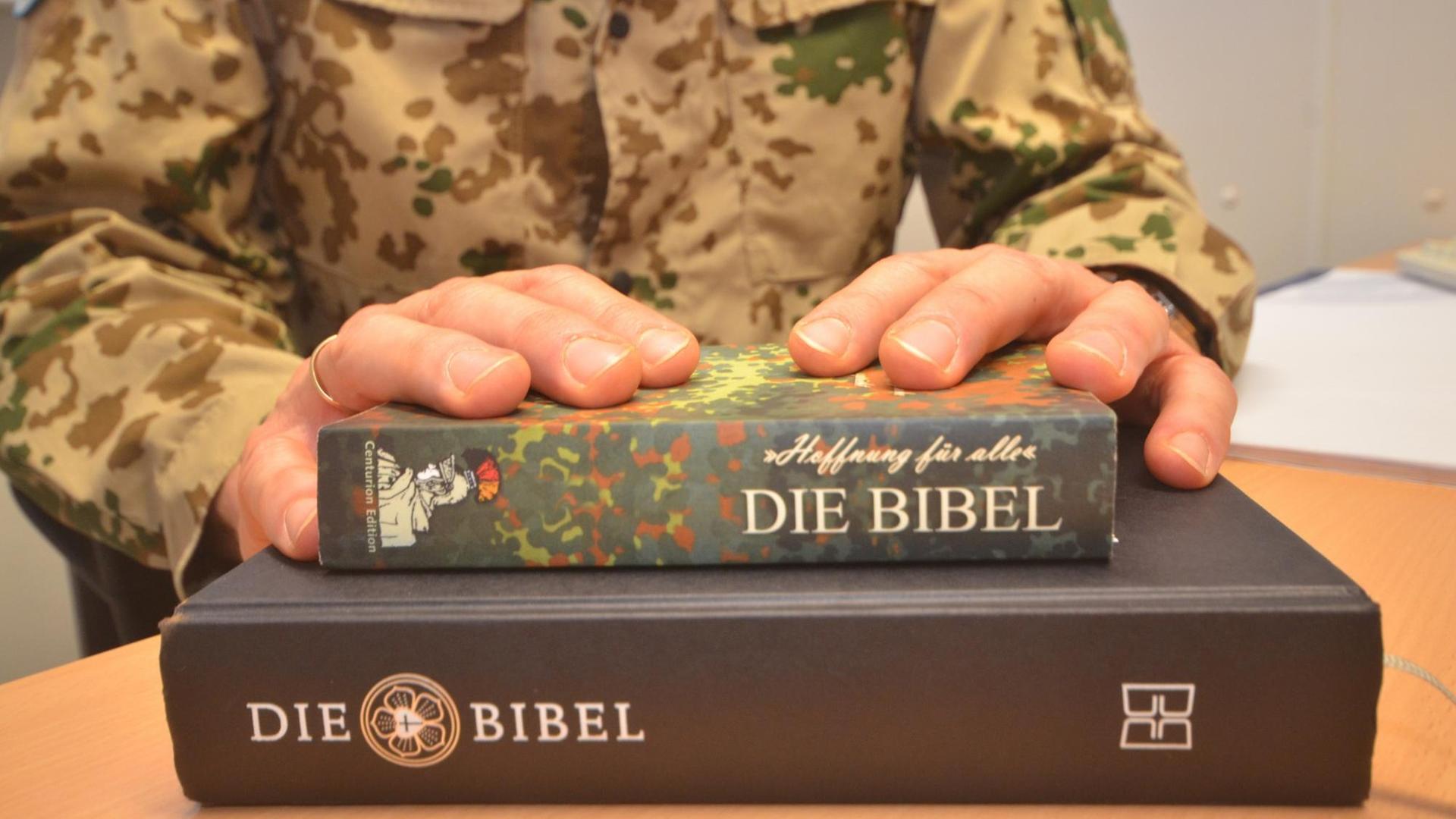 Eine Bibel in Flecktarn liegt auf einer klassisch gebundenen Bibel - die Hände des Militärpfarrers darüber.